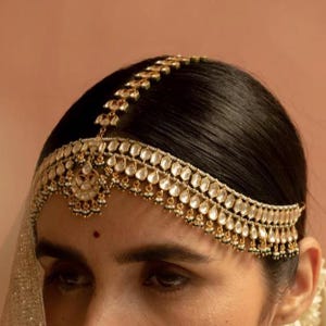 Op de afbeelding: Een goudkleurig hoofddeksel met heldere, druppelvormige stenen en kleine witte parels. Het sierlijke sieraad wordt op donker haar gedragen, met een kleine rode stip op het voorhoofd. Waarschijnlijk voor een speciale gelegenheid.