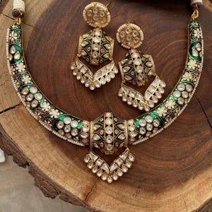 May include: An ornate necklace and earring set. The necklace features a curved design with green enamel, white floral accents, and clear gemstones. The matching earrings have a similar design. The jewelry is gold-toned and set on a wooden surface.
