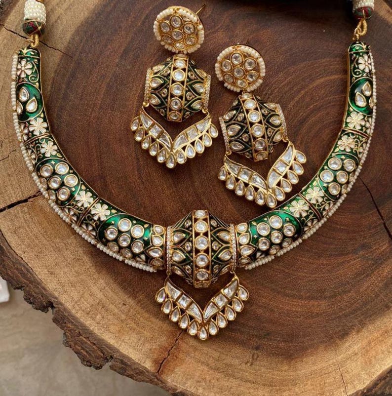 May include: An ornate necklace and earring set. The necklace features a curved design with green enamel, white floral accents, and clear gemstones. The matching earrings have a similar design. The jewelry is gold-toned and set on a wooden surface.