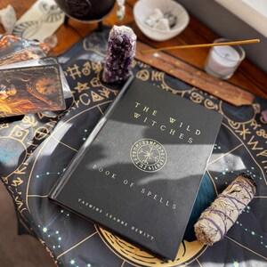 May include: A still-life arrangement featuring a black book titled "The Wild Witches Book of Spells" by Yasmine Leanne Hewitt, a purple amethyst crystal, tarot cards, a small cauldron, and a bundle of sage on a patterned cloth. A candle and incense are also present.