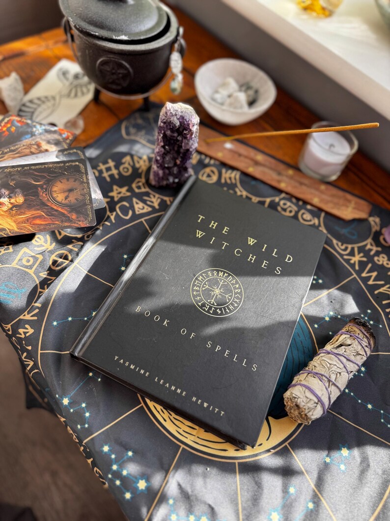 May include: A still-life arrangement featuring a black book titled "The Wild Witches Book of Spells" by Yasmine Leanne Hewitt, a purple amethyst crystal, tarot cards, a small cauldron, and a bundle of sage on a patterned cloth. A candle and incense are also present.