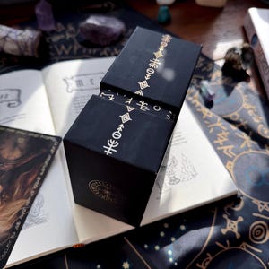 May include: Black box with gold-colored symbols and a lid, set on an open book with handwritten text. The box has a geometric design. The background includes a dark blue cloth with star patterns and other mystical items.