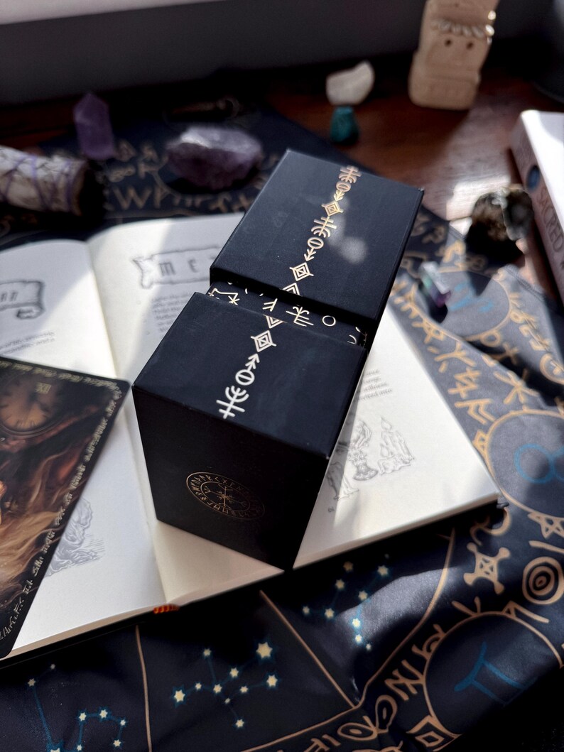 May include: Black box with gold-colored symbols and a lid, set on an open book with handwritten text. The box has a geometric design. The background includes a dark blue cloth with star patterns and other mystical items.