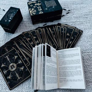 May include: A black tarot card deck with gold accents and a matching box. The cards are fanned out next to an open guidebook. The box has gold symbols and text. The cards and box are on a textured, light gray surface.