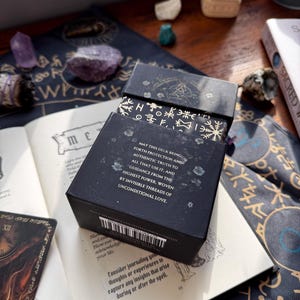 May include: A black tarot card box with gold accents and text, open on a book with a card visible. The box reads "MAY THIS DECK BRING FORTH PROTECTION AND AUTHENTIC TRUTH..." with a barcode on the side. Crystals and a smudge stick are also visible.