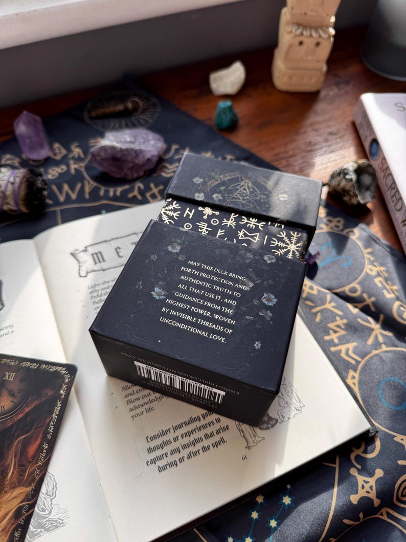 May include: A black tarot card box with gold accents and text, open on a book with a card visible. The box reads "MAY THIS DECK BRING FORTH PROTECTION AND AUTHENTIC TRUTH..." with a barcode on the side. Crystals and a smudge stick are also visible.