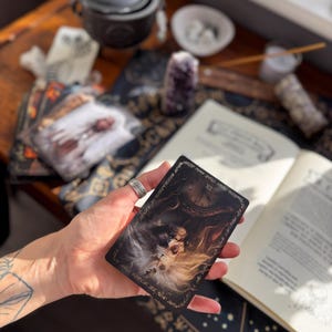 May include: A tarot card with a detailed illustration of a woman with long hair, a clock, and ornate borders. The card is held in a hand, with other mystical items like books and crystals in the background.