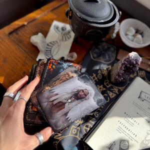 May include: A collection of tarot cards featuring fantasy art, a black cauldron, crystals, and a book with the text "MATERIALS" and "PREPARATION" on a wooden table. The cards depict images of angels and other mystical figures.