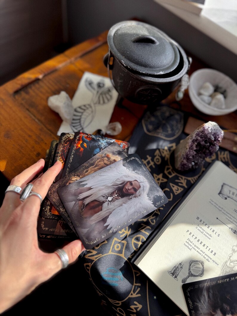 May include: A collection of tarot cards featuring fantasy art, a black cauldron, crystals, and a book with the text "MATERIALS" and "PREPARATION" on a wooden table. The cards depict images of angels and other mystical figures.