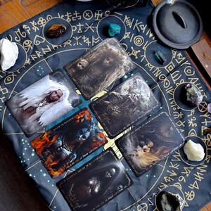May include: A collection of tarot cards with fantasy art, arranged on a dark blue mat with gold symbols. The cards feature images of mystical figures and scenes. Various crystals and a small cauldron are also present, creating a spiritual theme.