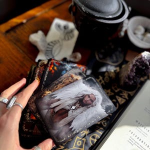 May include: A collection of tarot cards featuring fantasy art, with a card depicting a woman with white wings. The cards are on a table with a black cauldron, crystals, and a book with the word "PREPARATION".