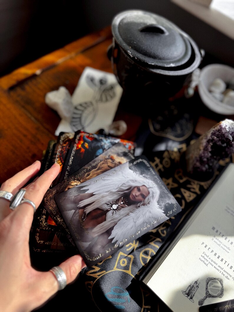 May include: A collection of tarot cards featuring fantasy art, with a card depicting a woman with white wings. The cards are on a table with a black cauldron, crystals, and a book with the word "PREPARATION".