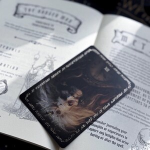 May include: A black tarot card with an illustration of a face and the words "The Hanged Man" in white script. The card is on top of an open book with text and illustrations. The book is open to page 61.