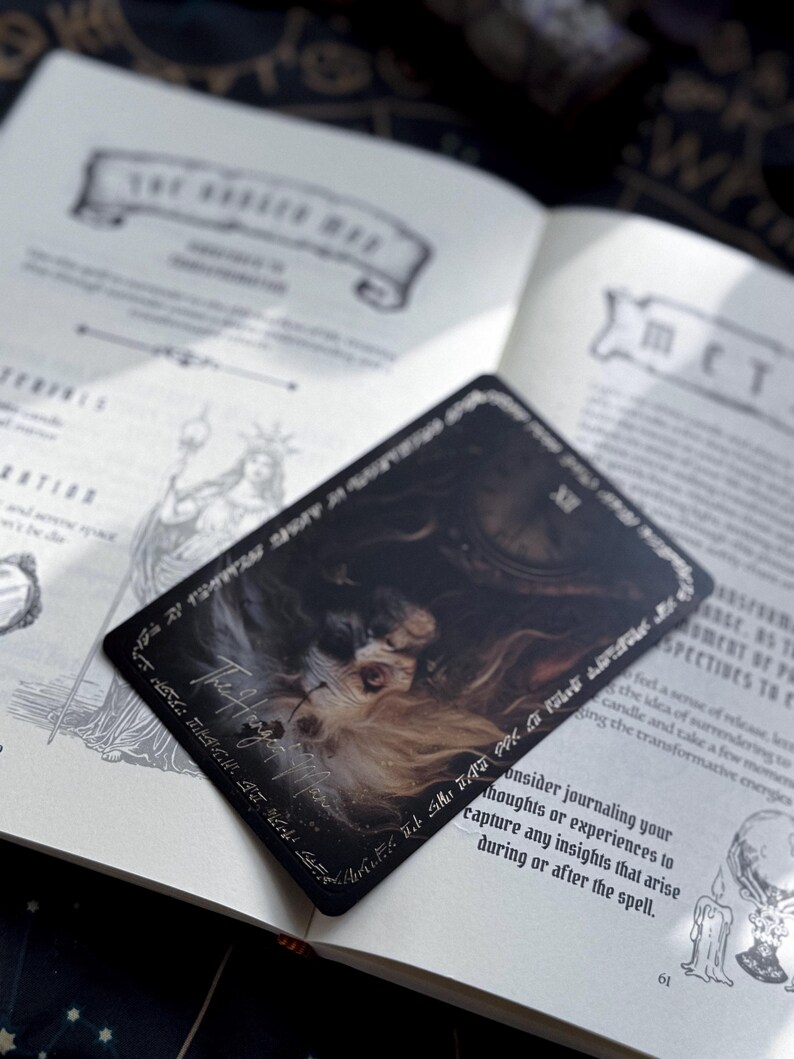 May include: A black tarot card with an illustration of a face and the words "The Hanged Man" in white script. The card is on top of an open book with text and illustrations. The book is open to page 61.