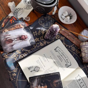 May include: An assortment of spiritual tools on a wooden surface. Includes a black cauldron, tarot cards, a book with the title "The Hanged Man", crystals, and a smudge stick. The cards feature angel imagery and the book has text.