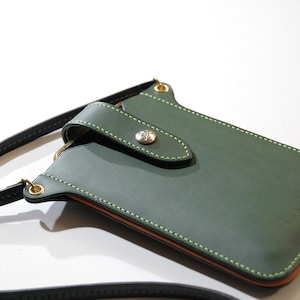 Könnte beinhalten: Ein grünes Leder-Handy-Etui mit Klappenverschluss und einem goldenen Knopf. Das Etui hat grüne Nähte und einen braunen Lederbesatz. Das Etui ist an einem schwarzen Lederband befestigt.