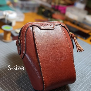May include: A small, reddish-brown leather bag with a rounded top and a zipper closure. The bag has a decorative tassel and the text "S-size" is visible on the side. The bag is stitched with a contrasting light-colored thread.
