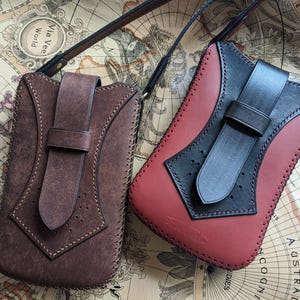 以下が含まれることがあります： ストラップ付きの革製の携帯電話ケース2つ。左側のケースは茶色でフラップ留め。右側のケースは赤で黒いフラップ留めです。