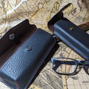 May include: Two black leather eyeglass cases with brown leather interiors and red stitching. The cases are open and have a snap closure. A pair of black eyeglasses are in the foreground.