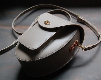 Peut inclure: Un sac à bandoulière en cuir gris avec une fermeture à rabat et une petite poche avant. Le sac a une longue bandoulière réglable et un fermoir en métal doré.