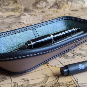 Könnte beinhalten: Ein braunes Leder-Stifttablett mit schwarzer Lederverkleidung und einem grauen Filzgrund. Zwei Stifte befinden sich im Tablett, einer schwarz und einer silberfarben.