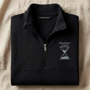 May include: Black quarter-zip sweatshirt featuring the text "abundance o'clock" and an hourglass design. The sweatshirt has a high collar and a silver zipper. The brand name "MERCER+METTLE" is printed on the collar.