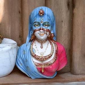 May include: A blue and pink painted ceramic bust of a woman wearing a veil and jewelry. The woman's face is partially obscured by a mask.