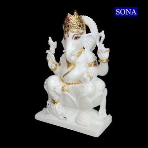 May include: White marble statue of Ganesha, the Hindu god of wisdom and prosperity, sitting on a lotus throne with a mouse at his feet. The statue is decorated with gold accents and red and green gemstones.