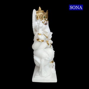 May include: White marble statue of Ganesha, the Hindu god of wisdom and prosperity, with gold accents. The statue is sitting on a white base and has a green gem on the forehead.