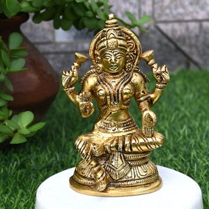 Pode incluir: Uma estátua de metal dourada da deusa hindu Lakshmi, sentada em um trono de lótus. Ela está segurando uma flor de lótus em cada mão e tem uma expressão serena no rosto.