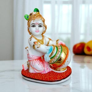 May include: A white and gold figurine of a seated deity, adorned with a crown and jewelry. The figure holds a gold and white pot with green and blue accents. It sits on a red patterned base. The figurine has a serene expression.