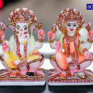 Può includere: Statue in marmo bianco delle divinità indù Ganesha e Lakshmi. Ganesha è raffigurato con una testa di elefante e quattro braccia, mentre Lakshmi è mostrata con quattro braccia e una corona. Entrambe le figure sono sedute su basi di fiori di loto e sono decorate con vernice colorata e accenti dorati.
