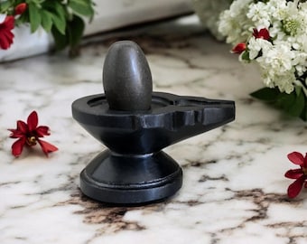 May include: A black stone Shiva lingam, a representation of the Hindu god Shiva, sits on a black stone base. The lingam is oval-shaped and has a smooth, polished surface. The base is intricately carved with a design that resembles a lotus flower.