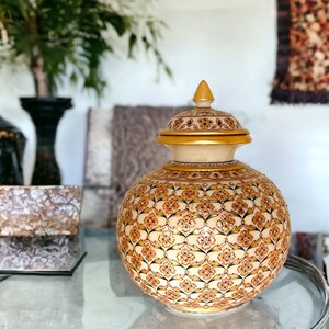 May include: A white ceramic jar with a gold lid and intricate floral patterns in gold, green, and pink. The jar is sitting on a glass table.