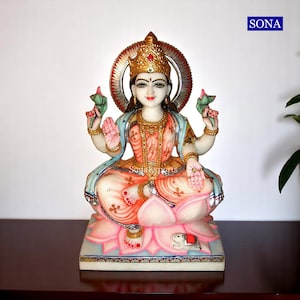 May include: A white marble statue of the Hindu goddess Lakshmi, sitting on a pink lotus flower. She is adorned with gold jewelry and a crown, and holds a lotus flower in each hand. The statue is painted with vibrant colors, including pink, blue, and gold.