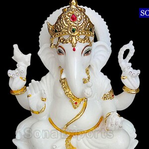 May include: White marble statue of Ganesha, the Hindu god of wisdom and prosperity. The statue is adorned with gold accents and red and green gemstones. The statue is sitting in a meditative pose with its trunk raised.