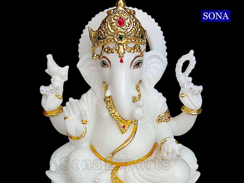 May include: White marble statue of Ganesha, the Hindu god of wisdom and prosperity. The statue is adorned with gold accents and red and green gemstones. The statue is sitting in a meditative pose with its trunk raised.