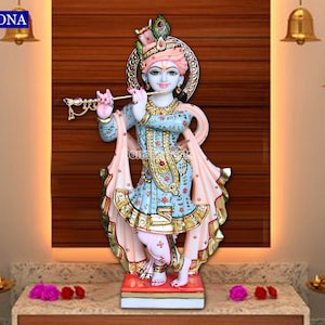 May include: A colorful marble statue of Krishna playing a flute. He is wearing a blue and gold patterned robe with a pink shawl. The statue is standing on a red and white base.