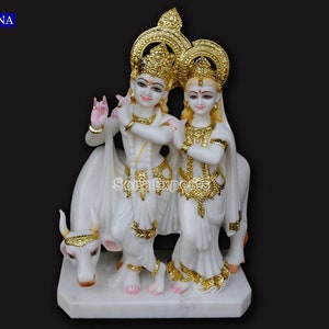 May include: White marble statue of Krishna and Radha standing together with a white cow. The statue is decorated with gold accents.