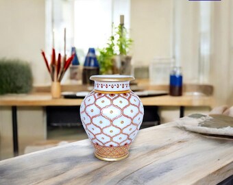 Könnte beinhalten: Weiße Marmorvase mit einem roten und goldenen geometrischen Muster. Die Vase ist rund und hat eine breite Öffnung. Sie steht auf einem Holztisch.