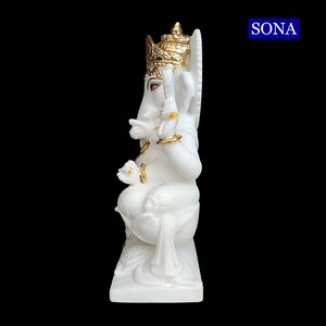 May include: White marble statue of Ganesha, the Hindu god of wisdom and prosperity, with gold accents. The statue is sitting on a base and has a crown on its head.