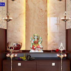 May include: A white marble statue of a Hindu goddess, Lakshmi, sitting on a lotus flower. The statue is placed on a white shelf in a home altar. The altar is decorated with two brass candle holders and a vase with red flowers.