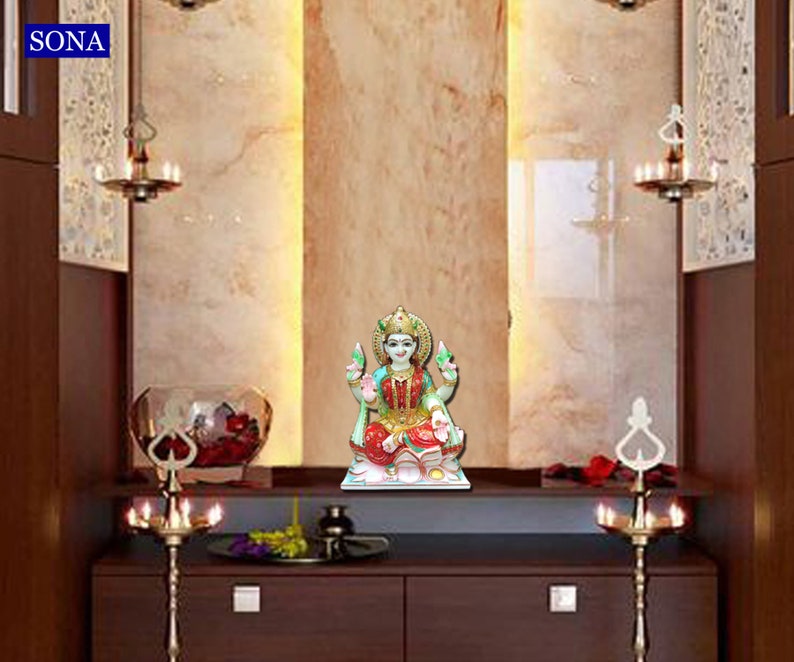 May include: A white marble statue of a Hindu goddess, Lakshmi, sitting on a lotus flower. The statue is placed on a white shelf in a home altar. The altar is decorated with two brass candle holders and a vase with red flowers.