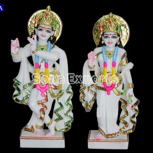 May include: White marble statue of Krishna and Radha, standing side by side. Krishna is playing a flute and Radha is holding a flower. Both figures are adorned with gold accents and colorful clothing.