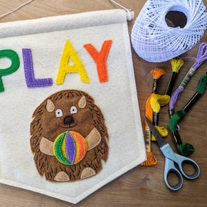 May include: A white felt banner with the word "PLAY" in rainbow colors. A brown felt hedgehog with a purple, orange, and green ball is below the word. The banner is hanging from a wooden dowel rod. There are also spools of embroidery floss in various colors and a pair of scissors.