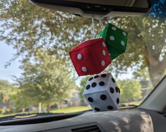 Op de afbeelding: Drie dobbelstenen auto-ornamenten hangen aan een achteruitkijkspiegel. De dobbelstenen zijn rood, groen en wit, met witte en zwarte stippen. De achtergrond is wazig, met bomen en een autodashboard.