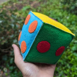 Può includere: Un giocattolo dadi in feltro con tessuto blu, verde, giallo, rosso e arancione. Il dado ha cerchi rossi e arancioni sul lato blu, cerchi rossi sul lato verde e tessuto giallo sulla parte superiore.