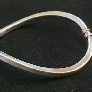 Peut inclure: Un bracelet jonc en métal argenté avec une finition lisse et polie, présentant une forme asymétrique rappelant un triangle. Le bracelet est doté d'un mécanisme d'ouverture à charnière sur un côté, permettant de l'ouvrir et de le fermer.