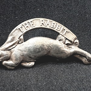 Puede incluir: Un broche de metal plateado que representa un conejo en movimiento. Encima del conejo hay una pancarta arqueada con las palabras "THE RABBIT". El broche está sobre un fondo oscuro y texturizado.