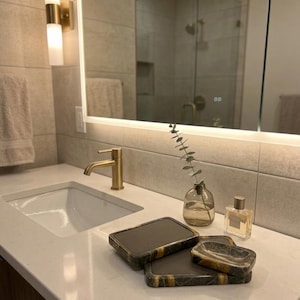 Op de afbeelding: Een moderne badkamer met een wit aanrecht, een gouden kraan en een rechthoekige wastafel. Decoratieve zeepbakjes met een donkergrijs en goud patroon zijn op het aanrecht geplaatst, samen met een kleine glazen vaas en een parfumflesje.